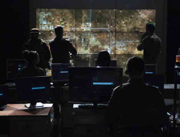 Military personnel watching drown footage on a large screen in a dark command center style room.