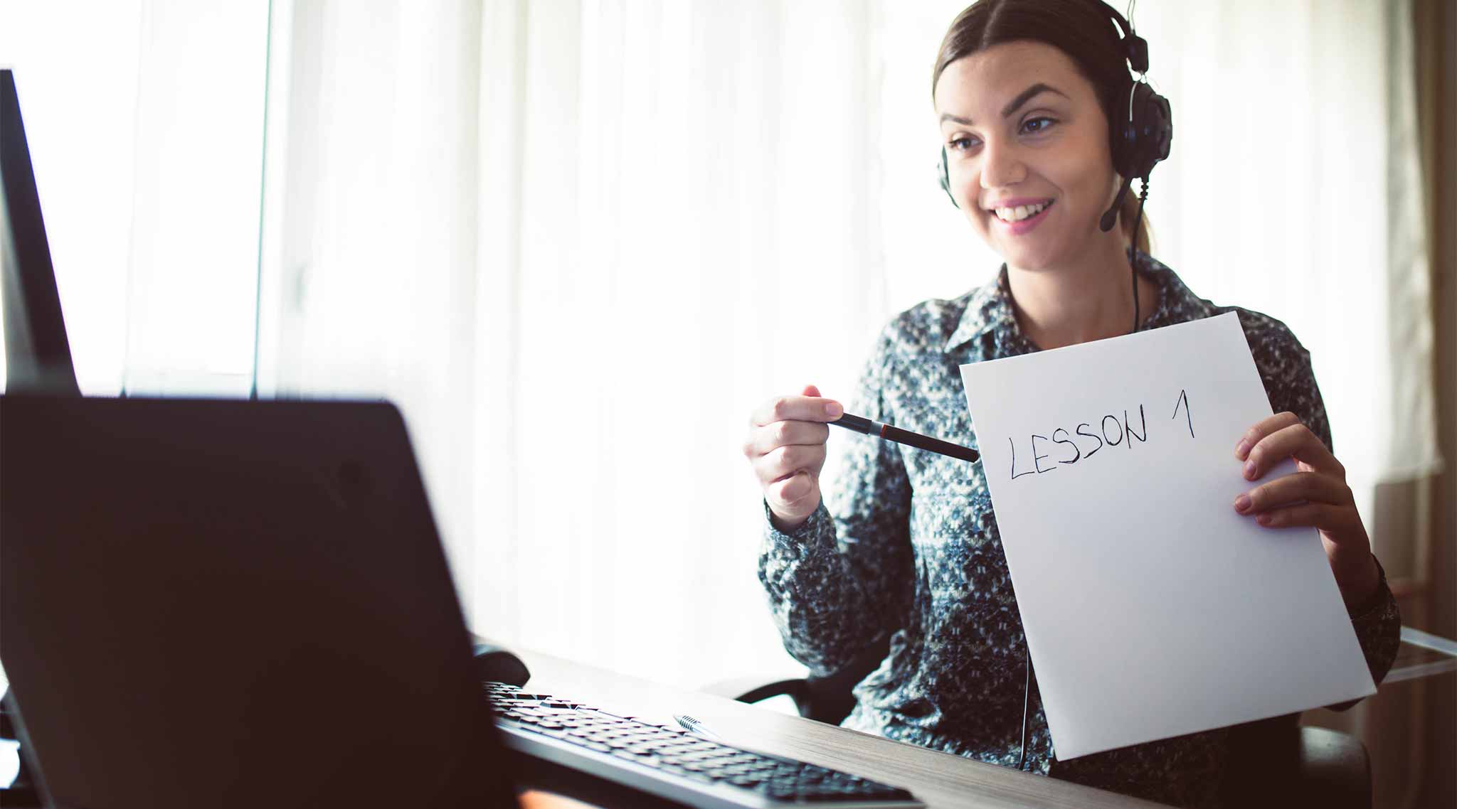 An online teacher talking to a laptop, holding up a paper that says "lesson 1."