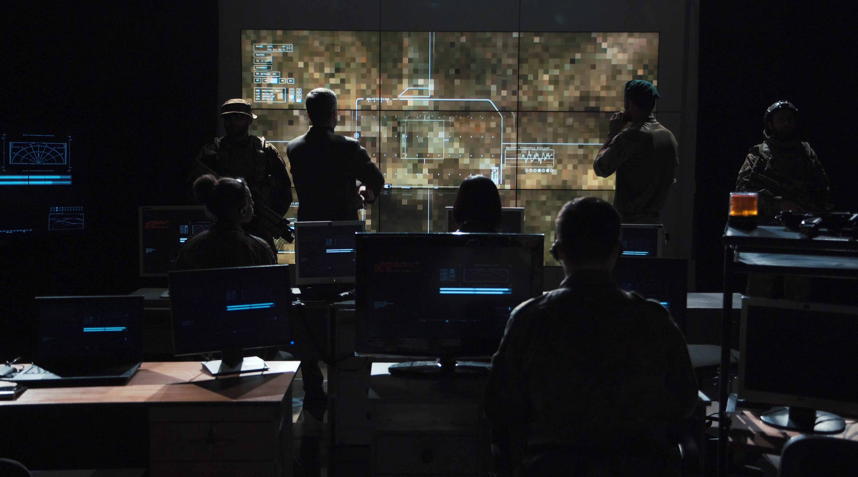 Military personnel watching drown footage on a large screen in a dark command center style room.