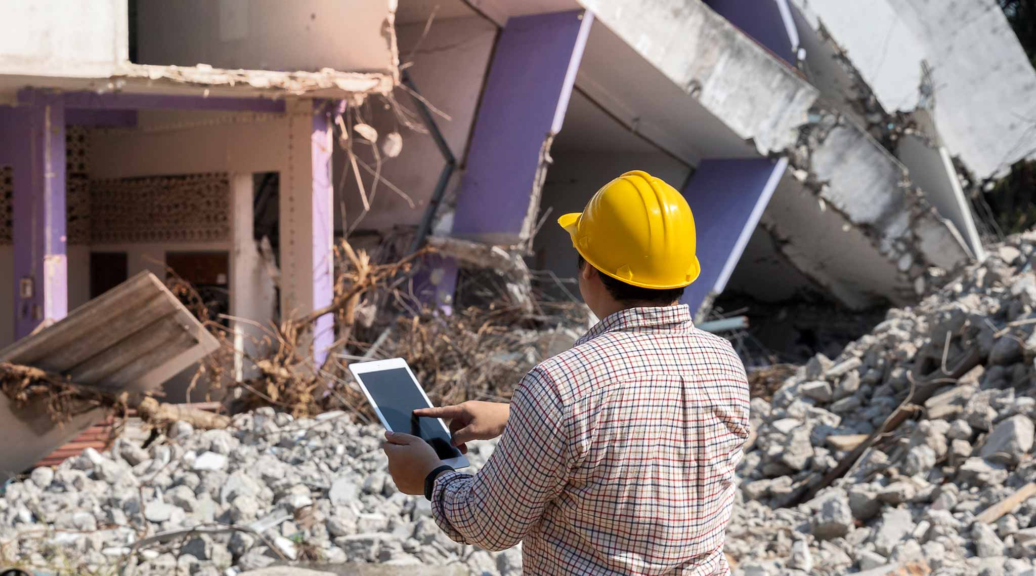Worker in a yellow hard hat standing in front of building rubble with a tablet.