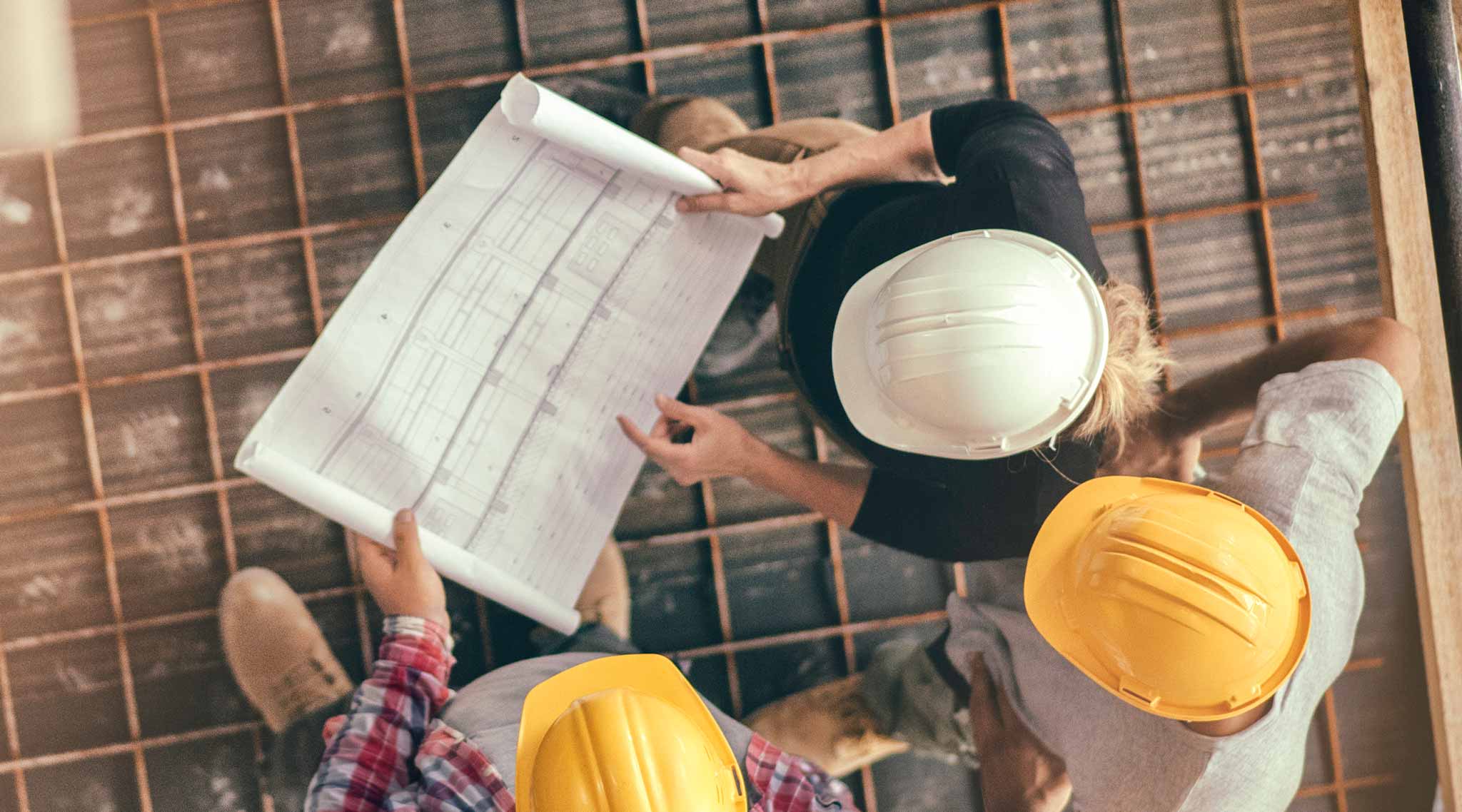 Overhead view of three people in hard hats holding blueprints standing on open rebar.