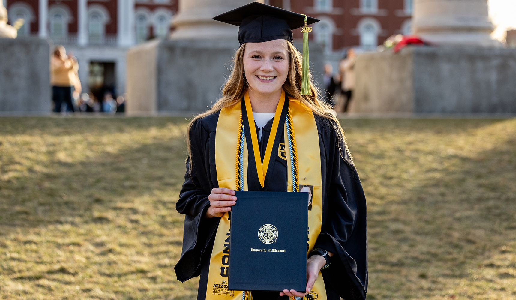 A student presenting her diploma to the camera, standing in front of the Columns.