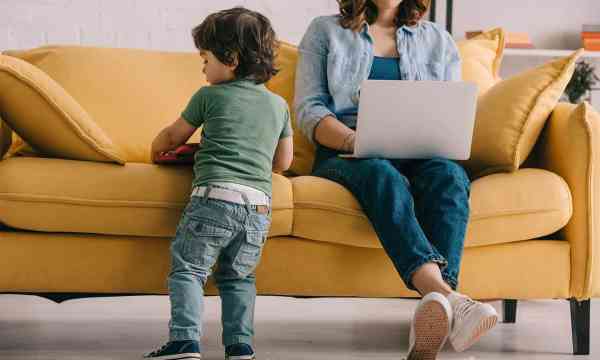 Kid standing near sofa while mother working with laptop stock photo.