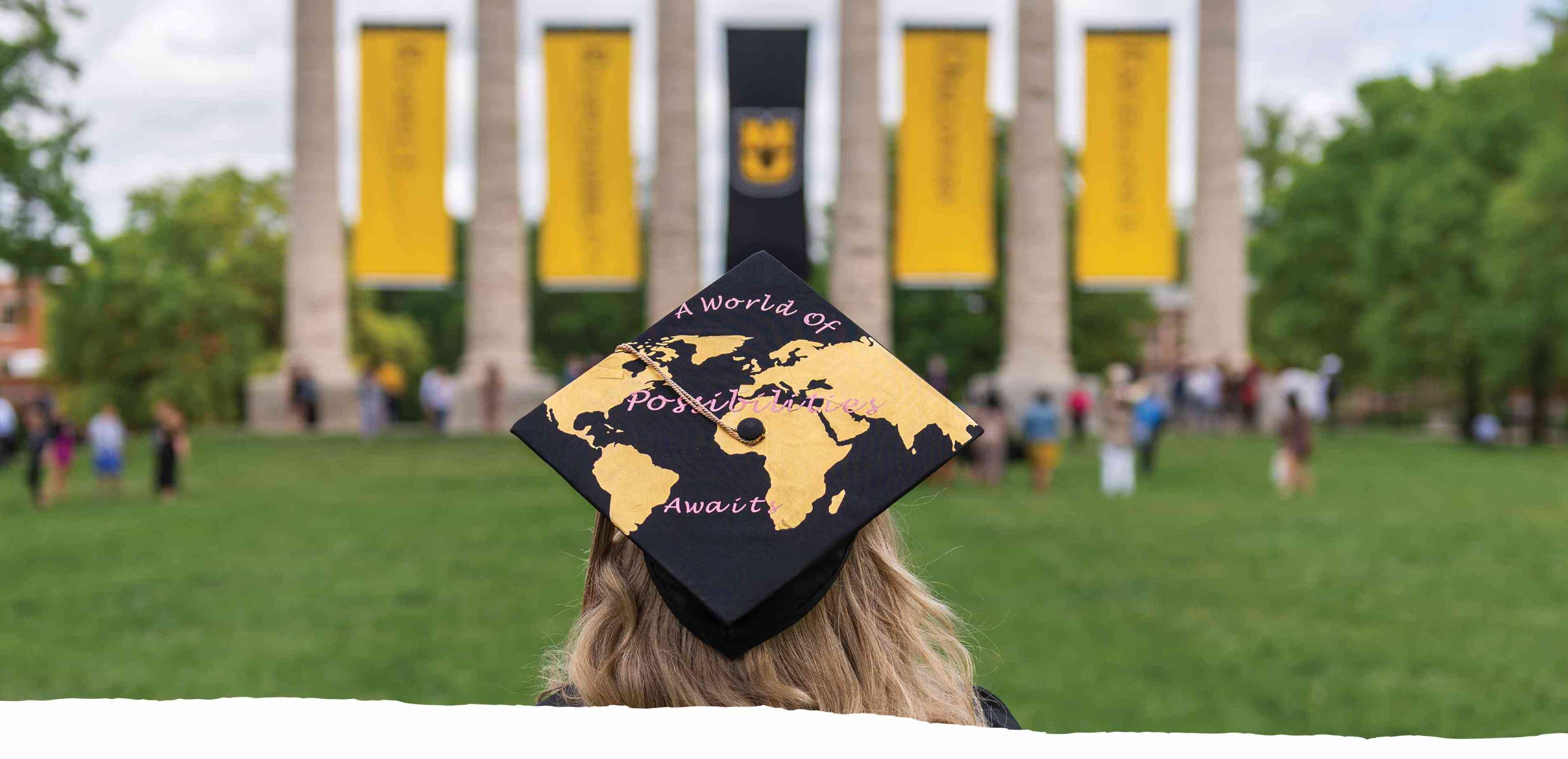 MU graduate facing columns on Mizzou's campus. Graduation cap reads "A World of Possibilities Awaits."