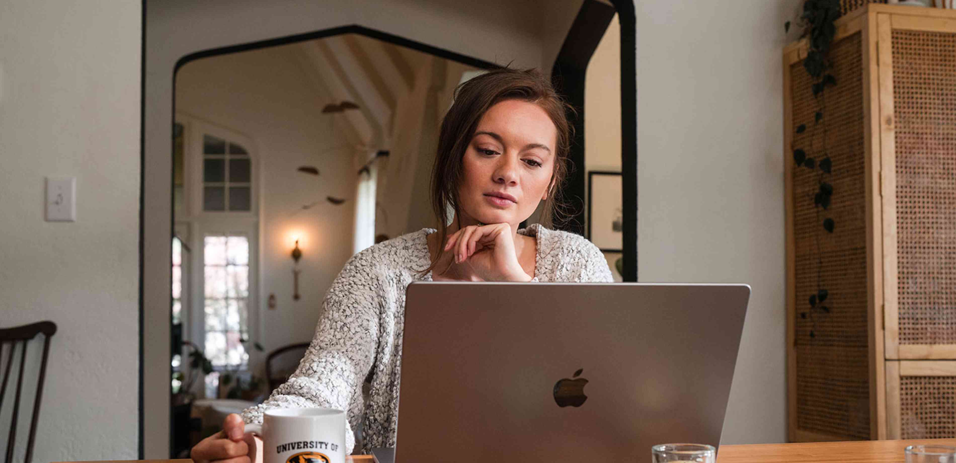 A woman on a laptop holding a Mizzou coffee mug.