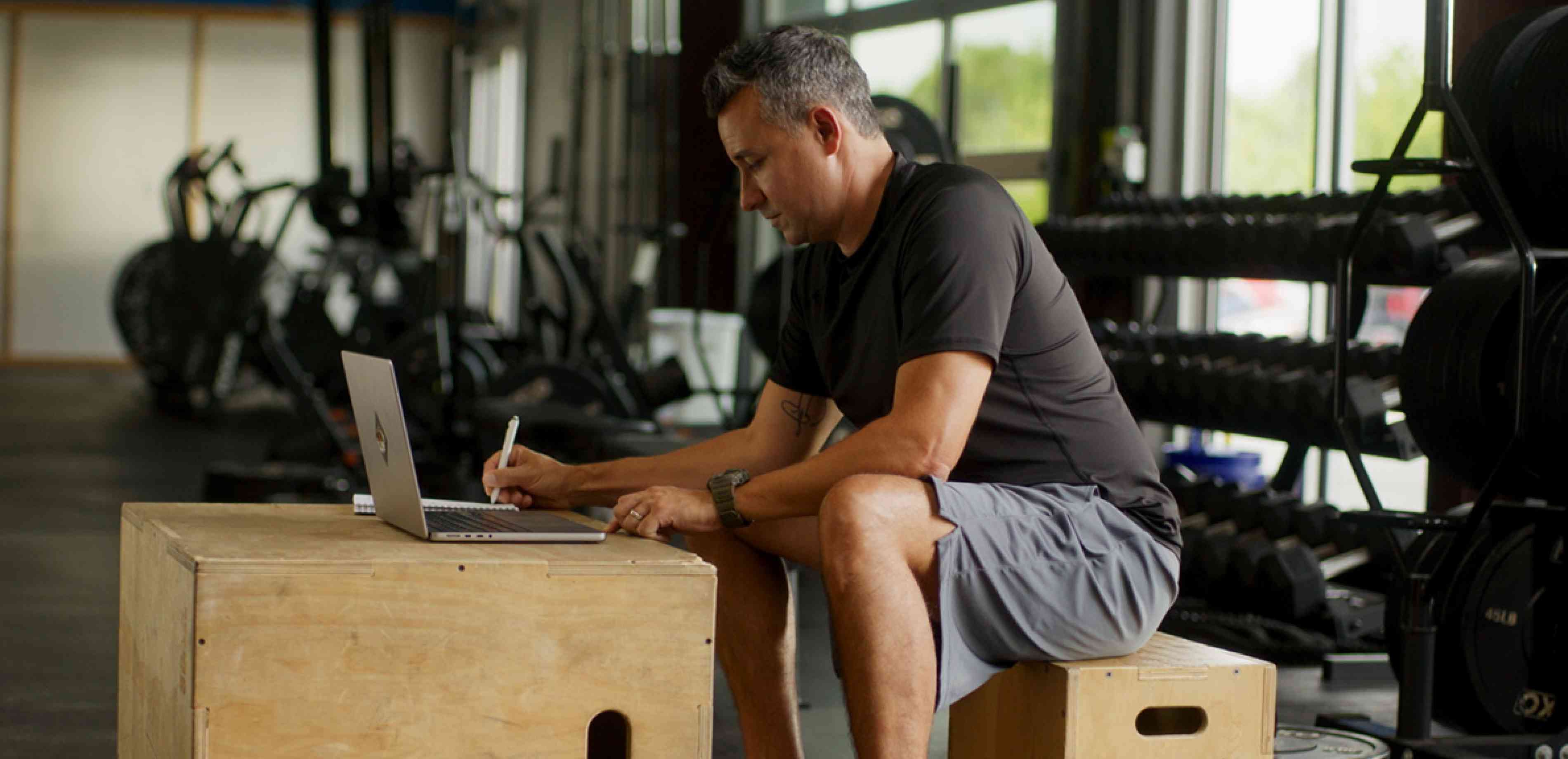 A man in a gym, sitting on a box, using a squat box as a table for his laptop, taking notes.