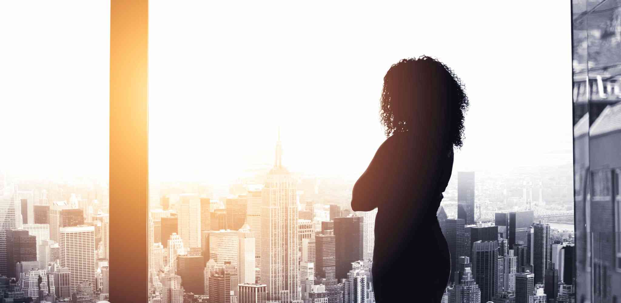 Women in silhouette looking out an upper floor office window at the sun setting over a city skyline.