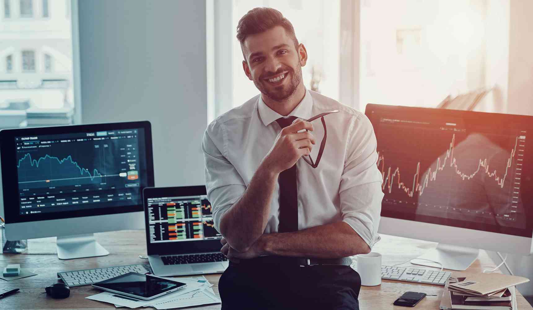 A finance professional sitting on a desk with computer screens behind them.