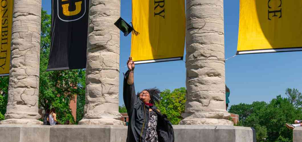 Kearsten Peoples, youth development MA, throws her graduation cap in the air in front of Mizzou's columns.