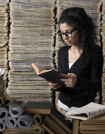 a female archivist studies a small book, surrounded by towers of newspapers