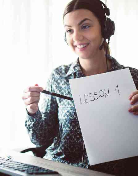An online teacher talking to a laptop, holding up a paper that says "lesson 1."