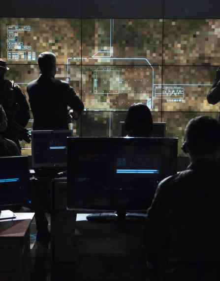 Military personnel watching drown footage on a large screen in a dark command center style room.