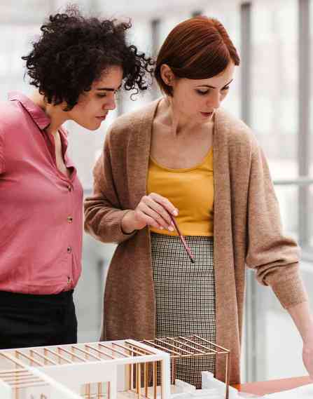 Two female young architects with model of a house standing in office, working and talking.