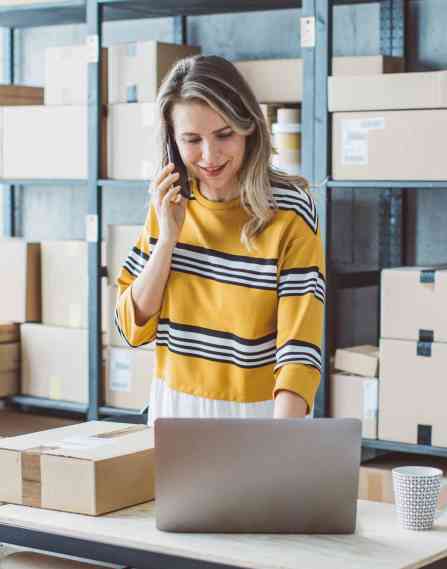 Digital merchandiser standing at a desk surrounded by boxes on the phone and looking at a laptop.