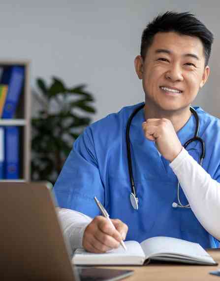 Male nurse wearing blue scrubs smiling at camera while working at a computer and taking notes on pen and paper.