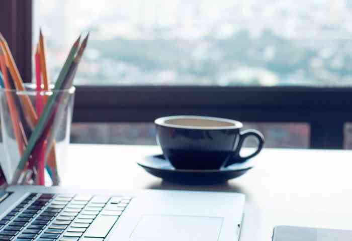 A desk with a laptop, cup of coffee, plant and cup of pencils on top of it.