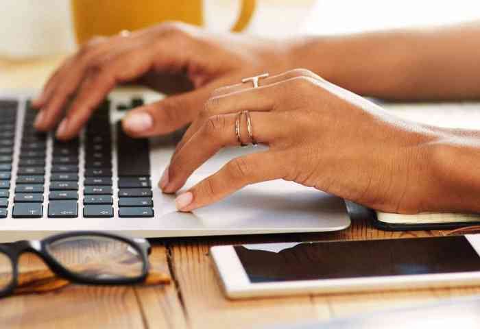 Someone typing on a laptop, phone and glasses next to them on the desk.