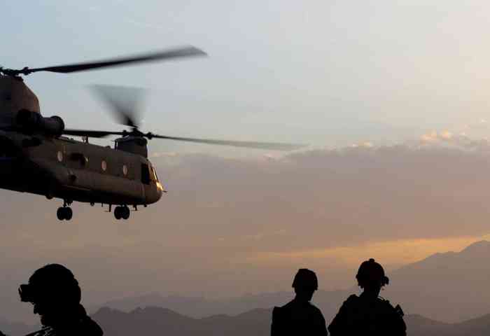 Military helicopter flying over troops in silhouette with the sun setting in the background.