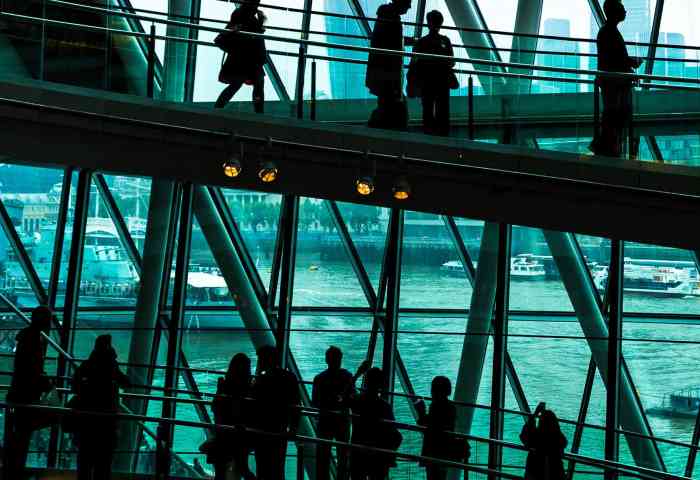 Crowds of people in silhouette and unrecognizable are walking down the modern spiral staircase. In the background we can see some of the skyscrapers that dominate the skyline of London.