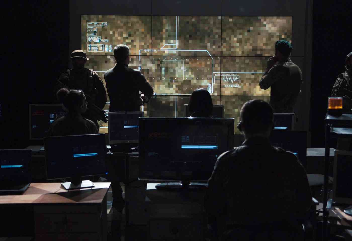 Military personnel watching drown footage on a large screen in a dark command center style room.