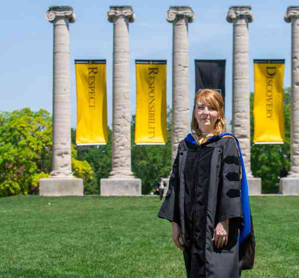 Bethanie Irons, PhD in art education, standing in front of the Mizzou columns wearing a doctoral gown.